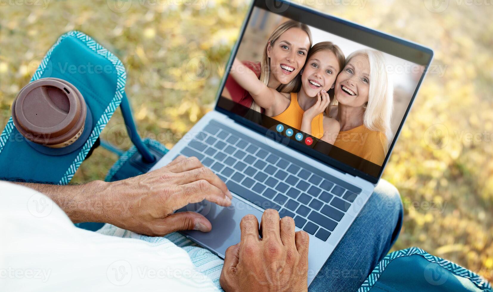A senior man sits on a blue lawn chair outdoors and uses a laptop to have a call with his family. He is smiling and appears happy to be connected to his loved ones photo