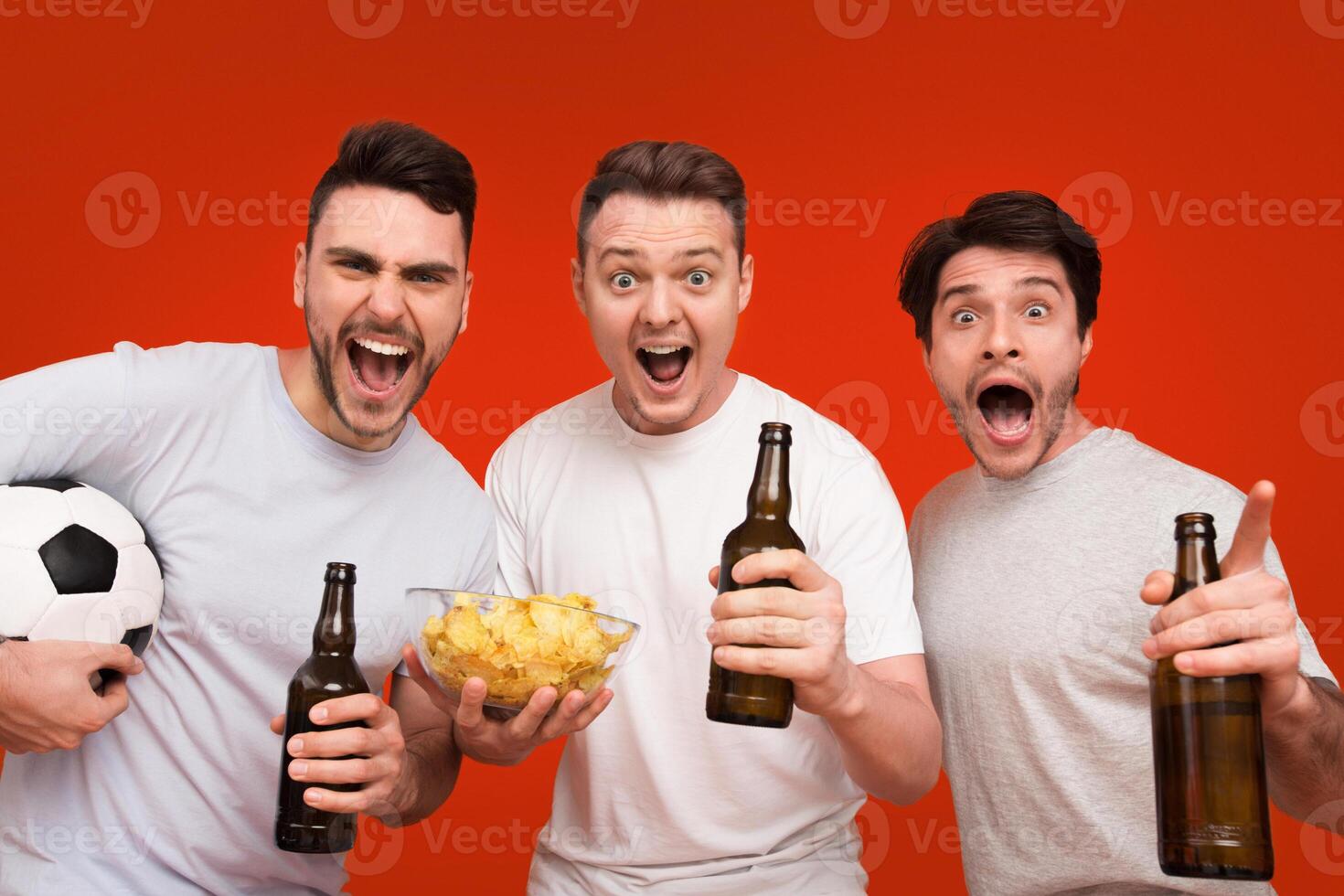 Three excited men holding a soccer ball, beer bottles, and chips bowl are cheering on a solid red background, depicting a sports celebration photo