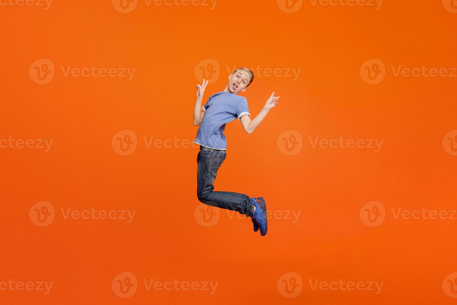Little ape. Boy grimacing, showing tongue and peace sign, jumping in air on orange studio background photo