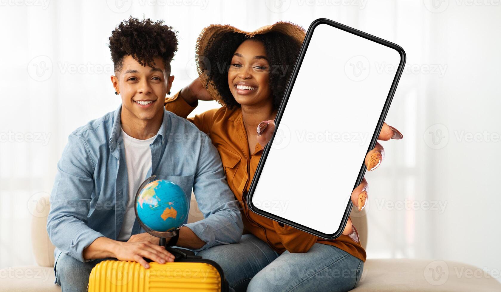 A young, smiling black couple sits closely together, holding a blank-screen smartphone suitable for mock-up displays, suggesting they are planning a trip or vacation. photo
