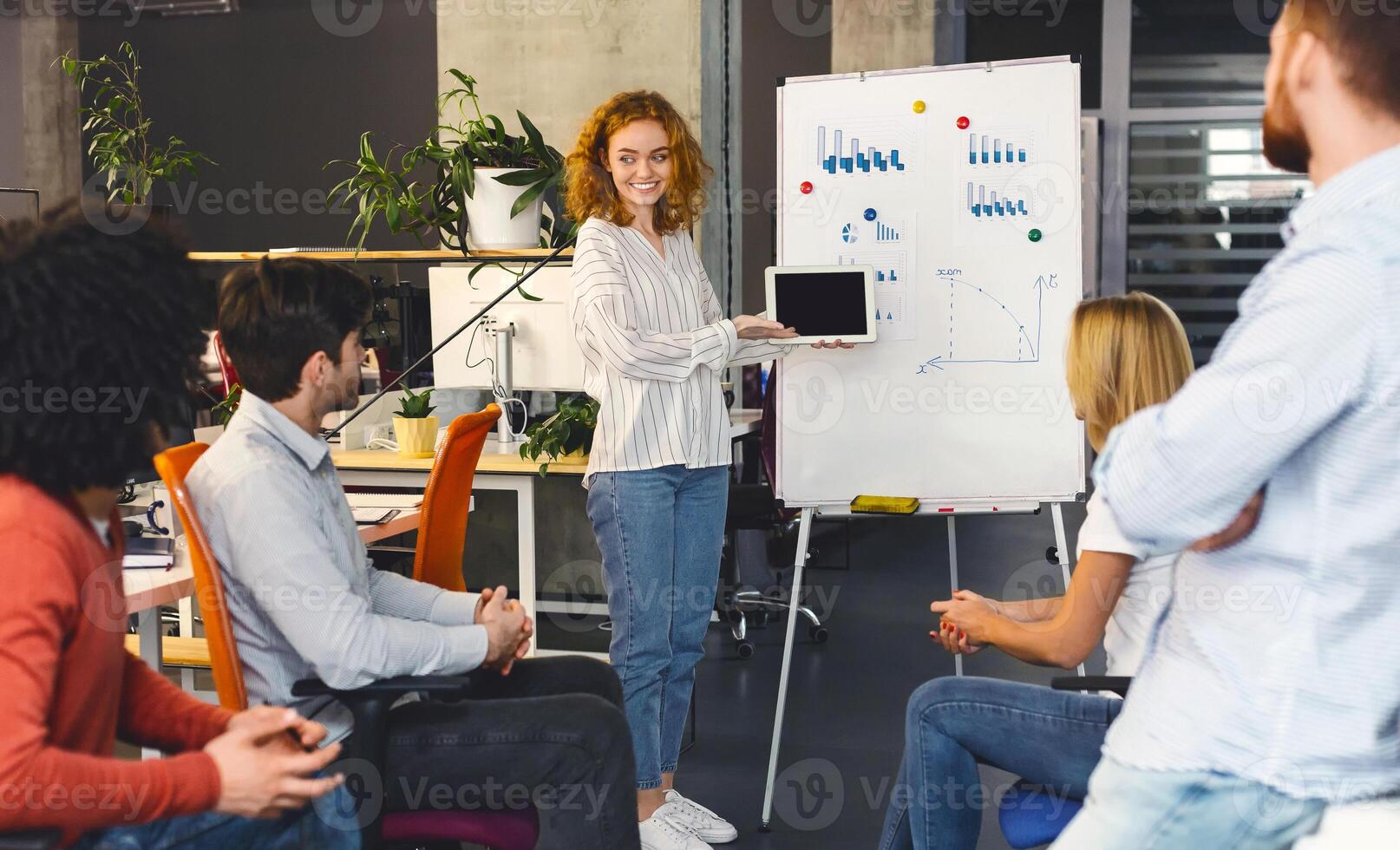 A young woman with curly hair is presenting to her colleagues in a modern office. She stands beside a whiteboard with charts and diagrams, smiling confidently as her team photo
