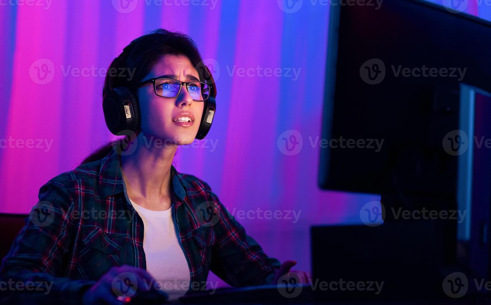 A young woman with glasses and headphones is seated in front of a computer screen. She appears to be focused intently on the screen, with a serious expression on her face photo