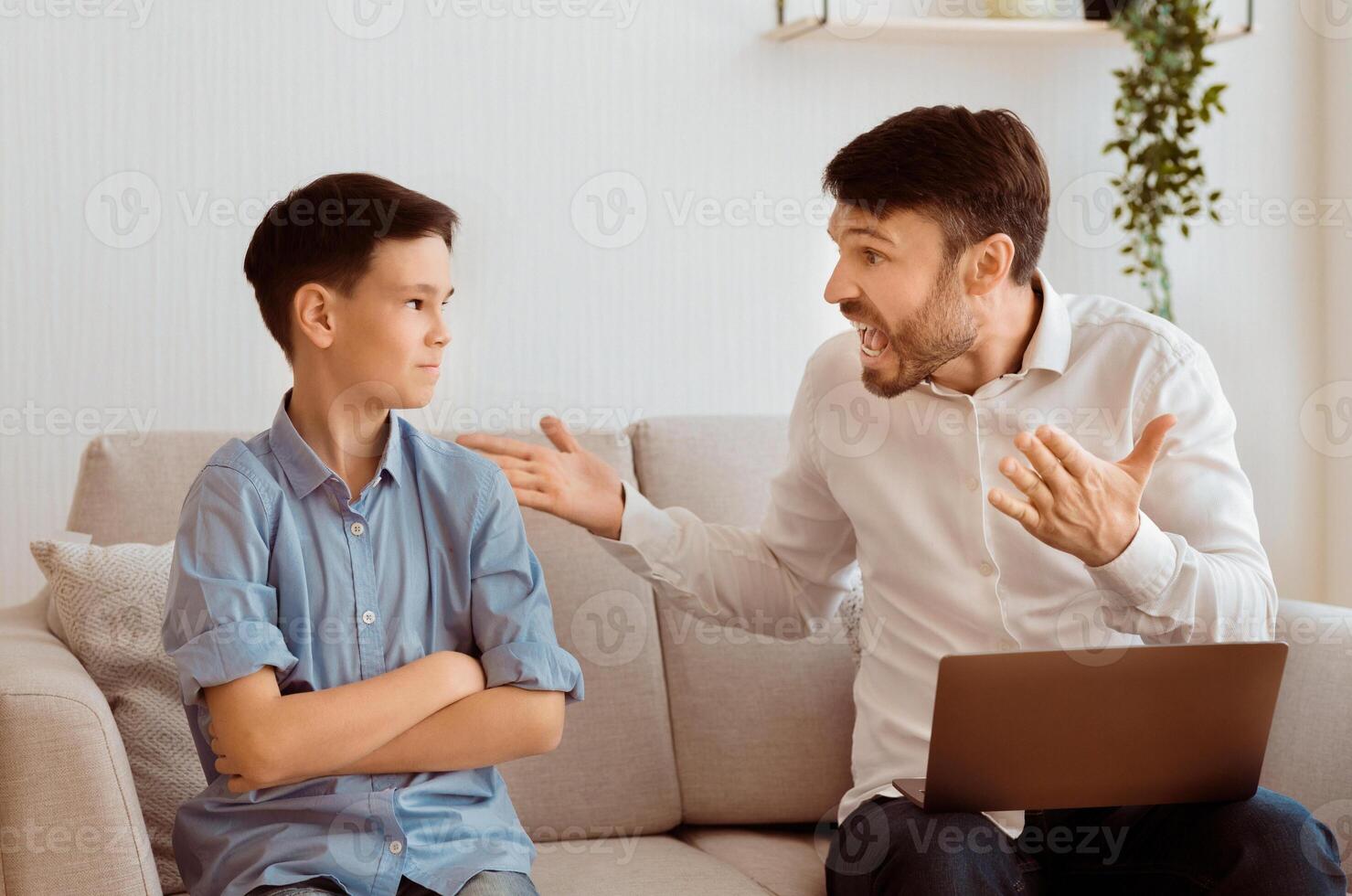 Aggressive Father. Angry Dad Yelling At Offended Son Working On Laptop Sitting On Couch Indoor. Selective Focus photo