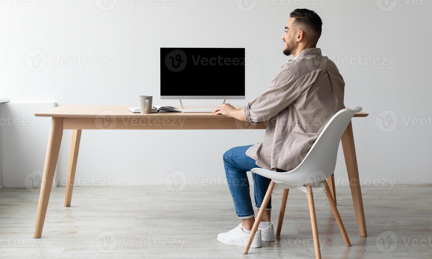 Rear Back View Of Happy Smiling Arab Man Looking At Pc Computer With Blank Black Screen For Mock Up Template Sitting At Desk At Home Office, Free Copy Space. People, Technology, Remote Work Concept photo