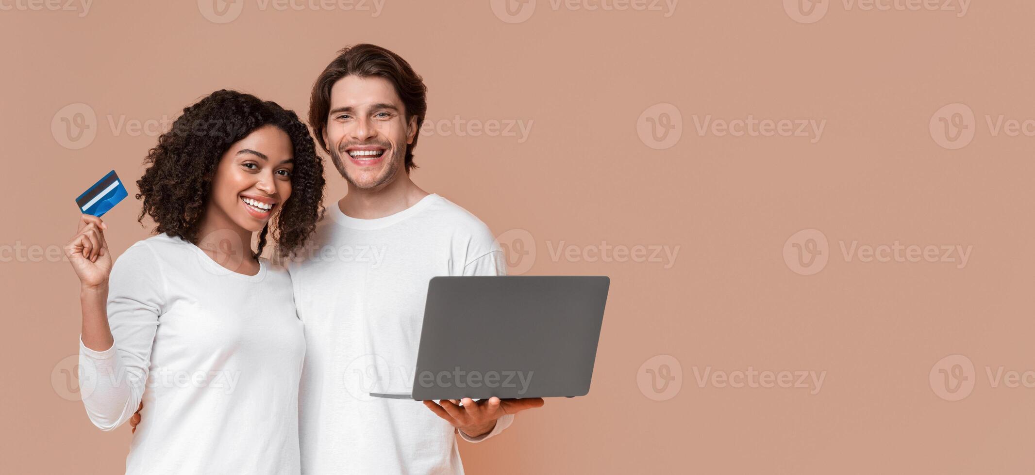 Easy Payments Concept. Happy multiracial spouses holding laptop and credit card, posing together over yellow studio background, copy space photo