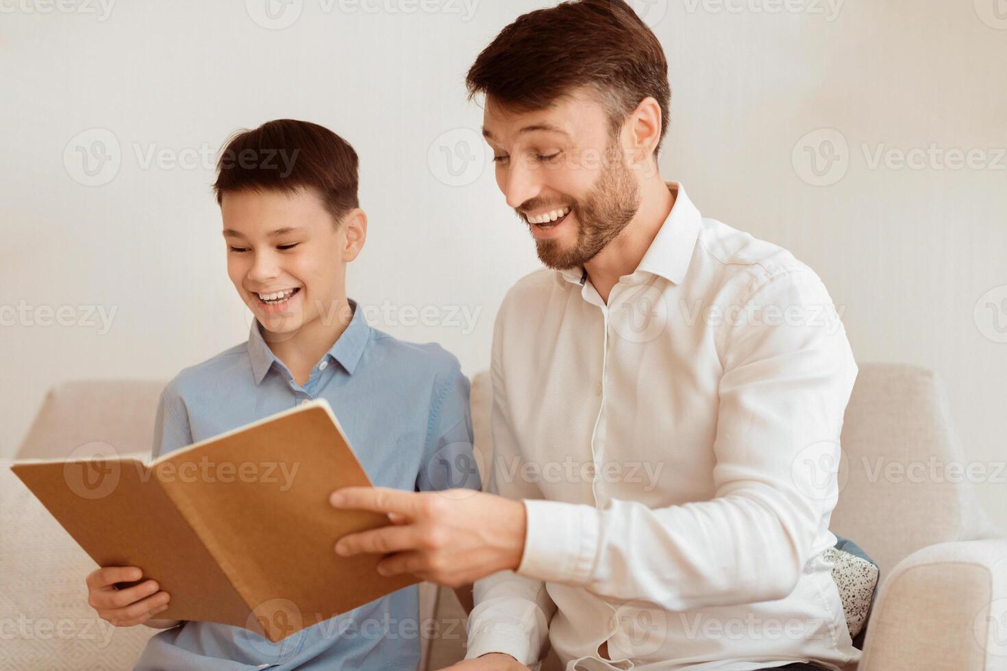 Happy Father And Son Reading Book And Laughing Sitting Together On Couch At Home. Parenthood And Family Relationship. Selective Focus photo
