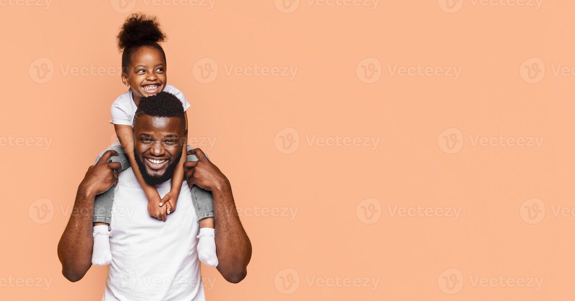 A happy black father and daughter pose for a picture against a peach-colored background. The little girl is sitting on her fathers shoulders, and both are smiling brightly at the camera, copy space photo