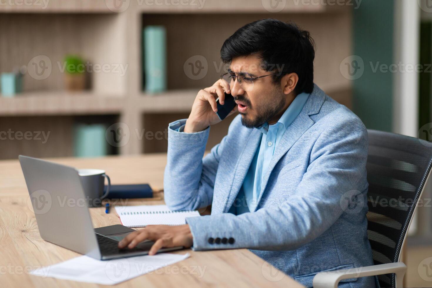 Indian businessman sits at a desk in a modern office, holding a phone to his ear with a frustrated expression on his face. He is looking directly at the phone, with one hand on his laptop keyboard photo