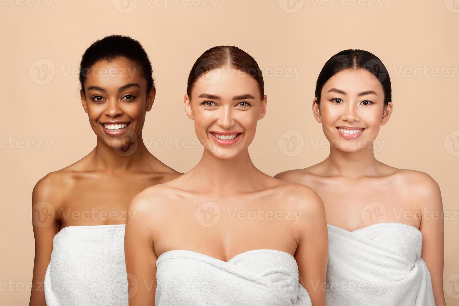 Three Happy Diverse Girls Wrapped In White Towels Posing Standing On Gray Studio Background, Smiling To Camera. Body Care Concept photo