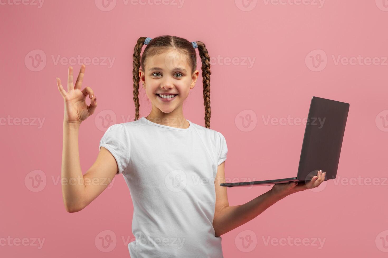 A young girl is standing against a pink background, holding a laptop computer in her hands. She appears to be engrossed in the screen, possibly playing a game or watching a photo