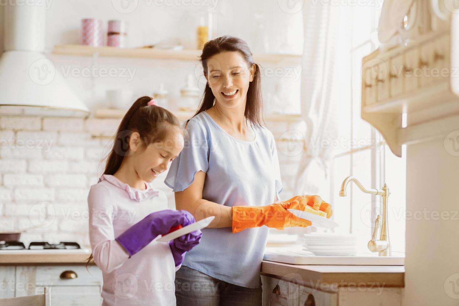 This image shows a mother and daughter washing dishes in a modern kitchen. Mother is wearing orange gloves and is washing a plate in the sink. Daughter is wearing purple gloves and is holding a plate photo