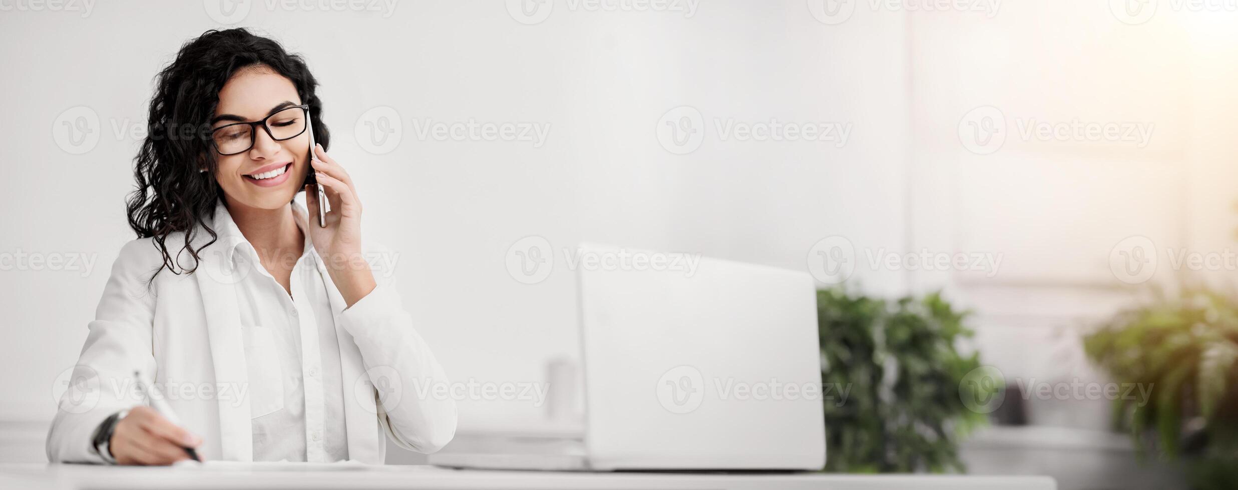 Business Call. Smiling mexican secretary talking on phone and writing notes at work, consulting client, copyspace photo
