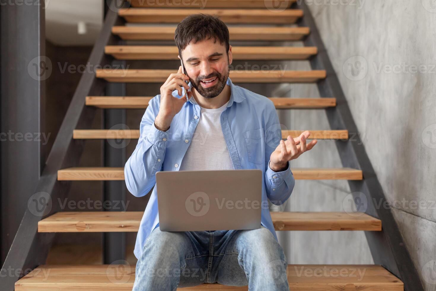 A man is sitting on wooden stairs in a home, talking on a cell phone while holding a laptop on his lap. He is gesticulating with one hand as he speaks, likely discussing something related to his work. photo