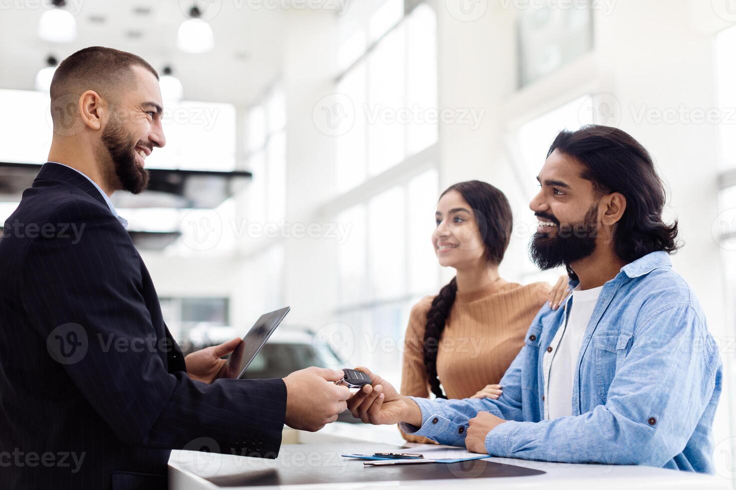 A joyful Indian couple stands at a dealership counter, receiving a new set of car keys from a professional salesman. The showroom is well-lit photo