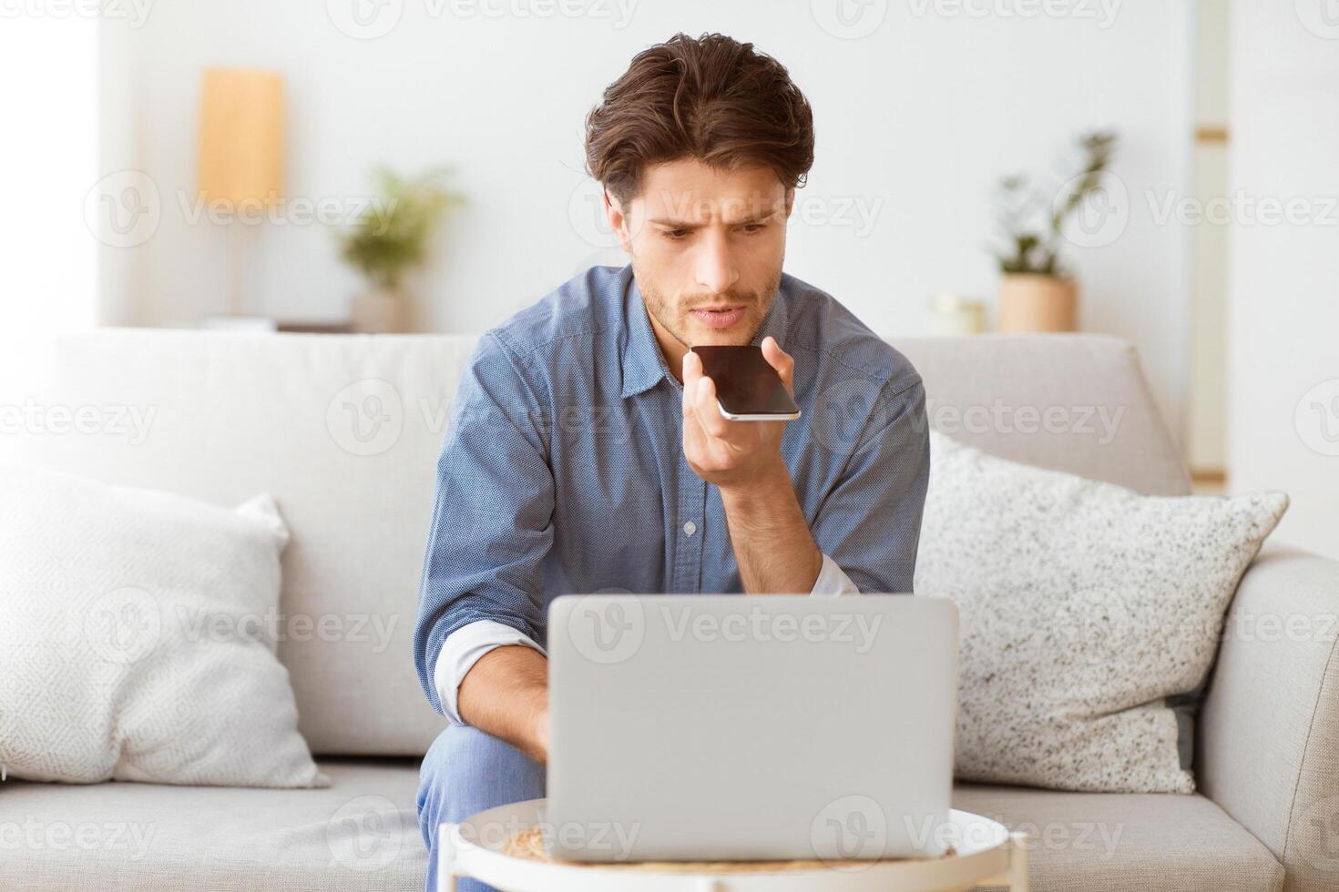 A man sits on a couch in his home and uses the voice assistant on his smartphone while working on a laptop. photo