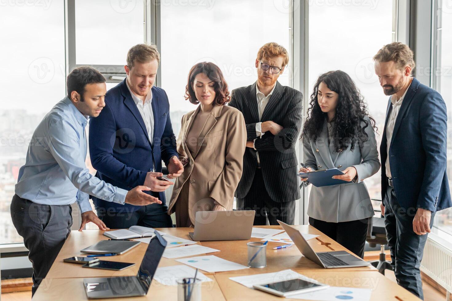 A group of business professionals gather around a conference table in a modern office setting. They are engaged in a discussion, reviewing documents and using laptops. photo