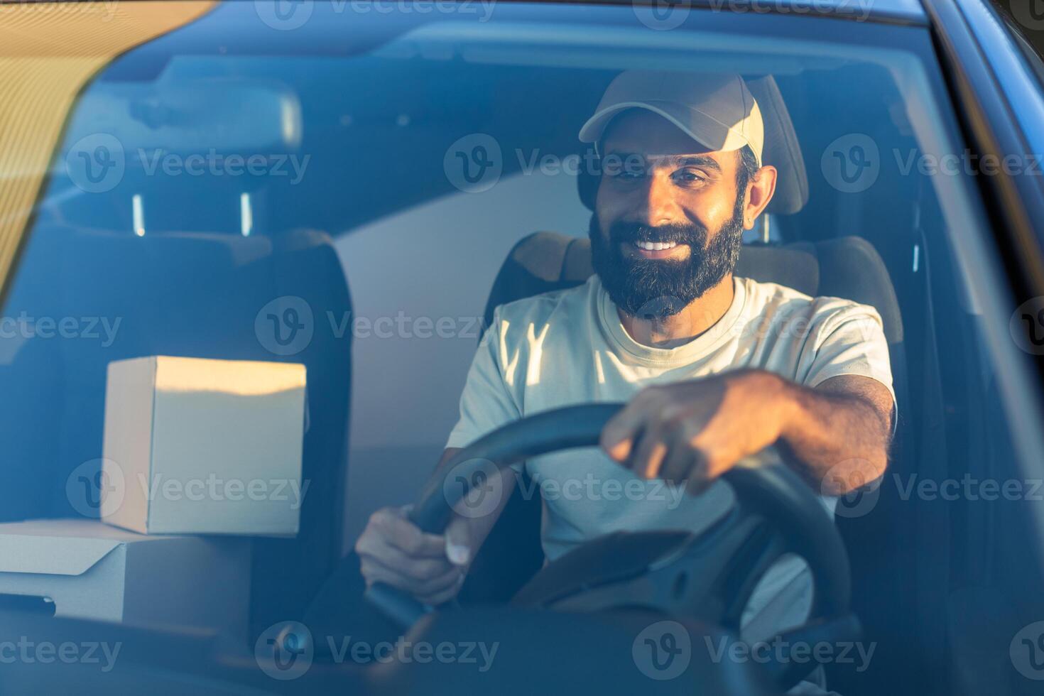 A cheerful Indian delivery driver navigates his vehicle through city streets early in the morning. He is seated behind the wheel, with packages visible beside him photo