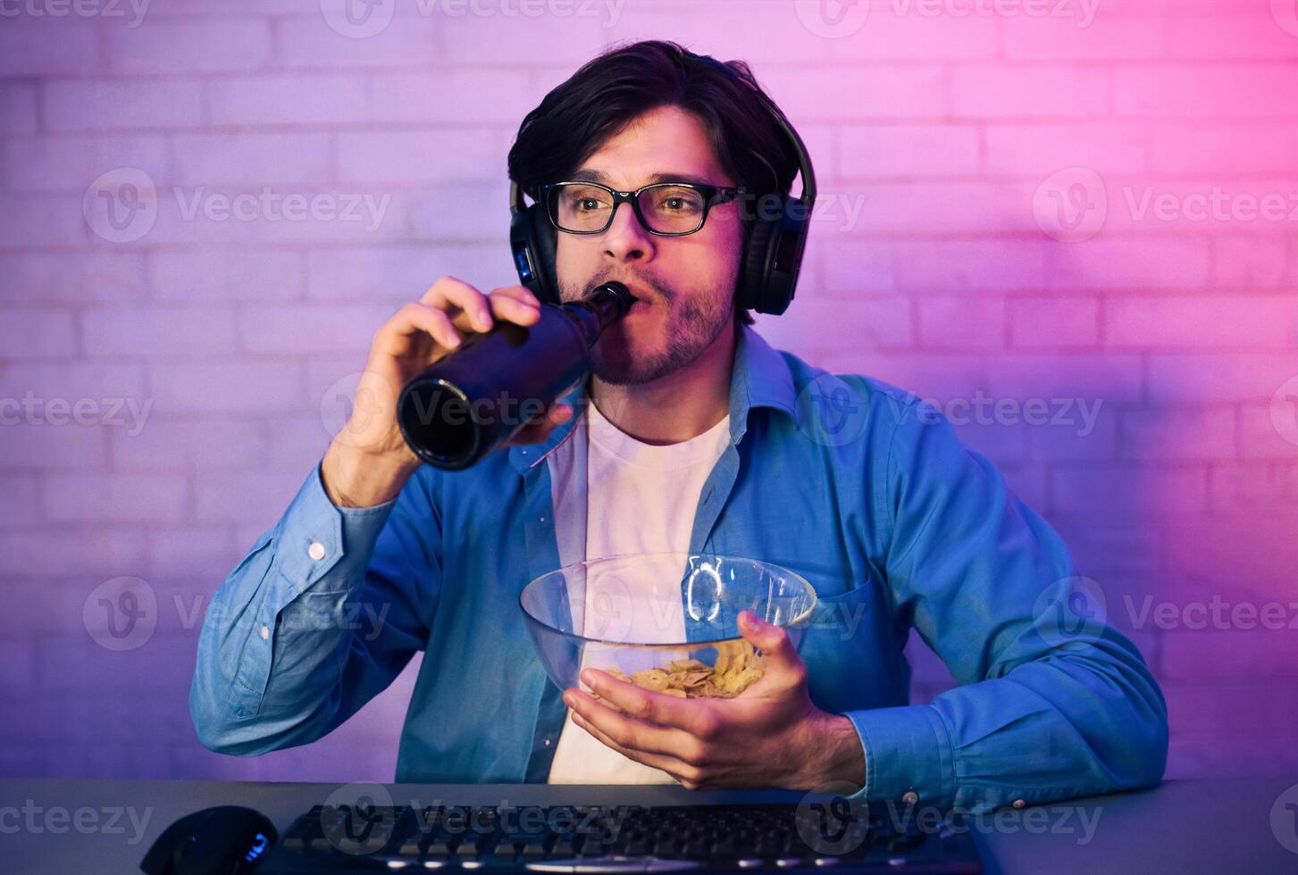 A young man wearing headphones and glasses is sitting at a desk, drinking from a black bottle, and eating chips from a bowl. He is likely playing a game as the desk has a keyboard and mouse. photo