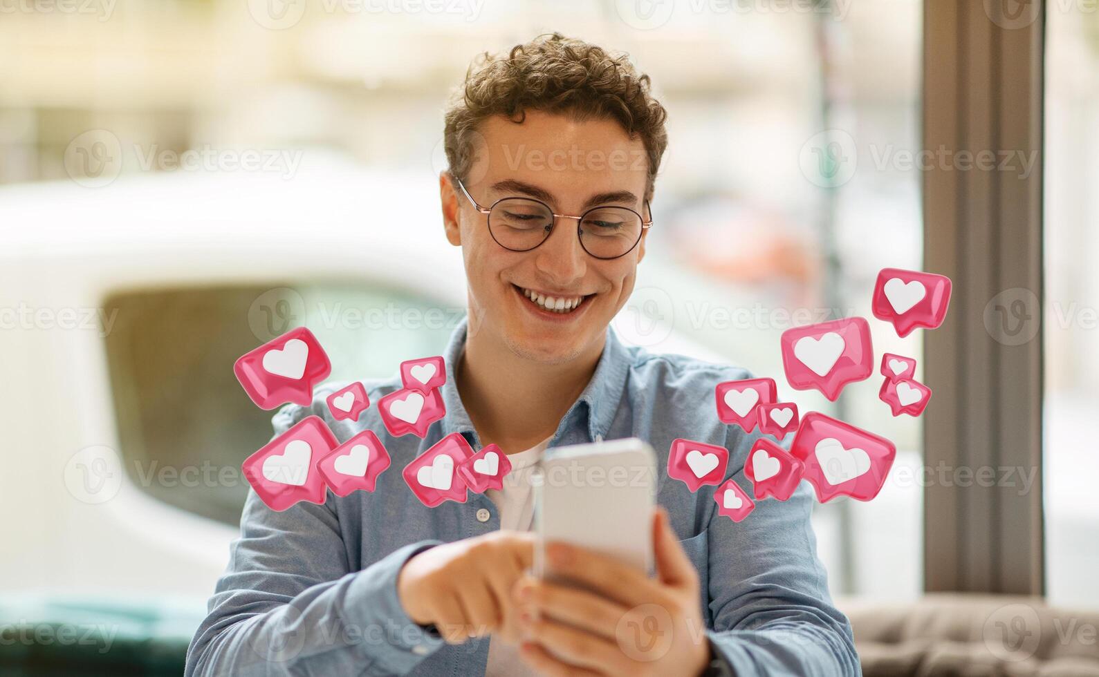 A man wearing glasses smiles broadly as he looks at his phone. A stream of social media heart icons, in a white heart inside a pink box, float around him. He is seated indoors, possibly in a cafe photo