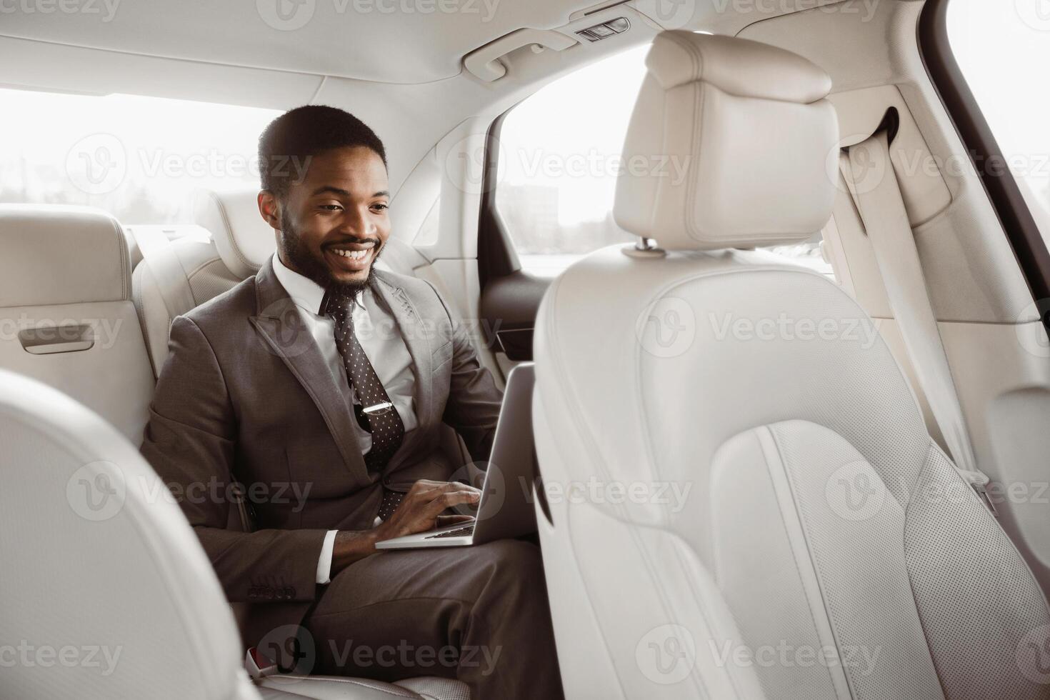 African American man in a business suit is seated in the back seat of a car, working on a laptop. He is smiling and appears to be focused on his work photo