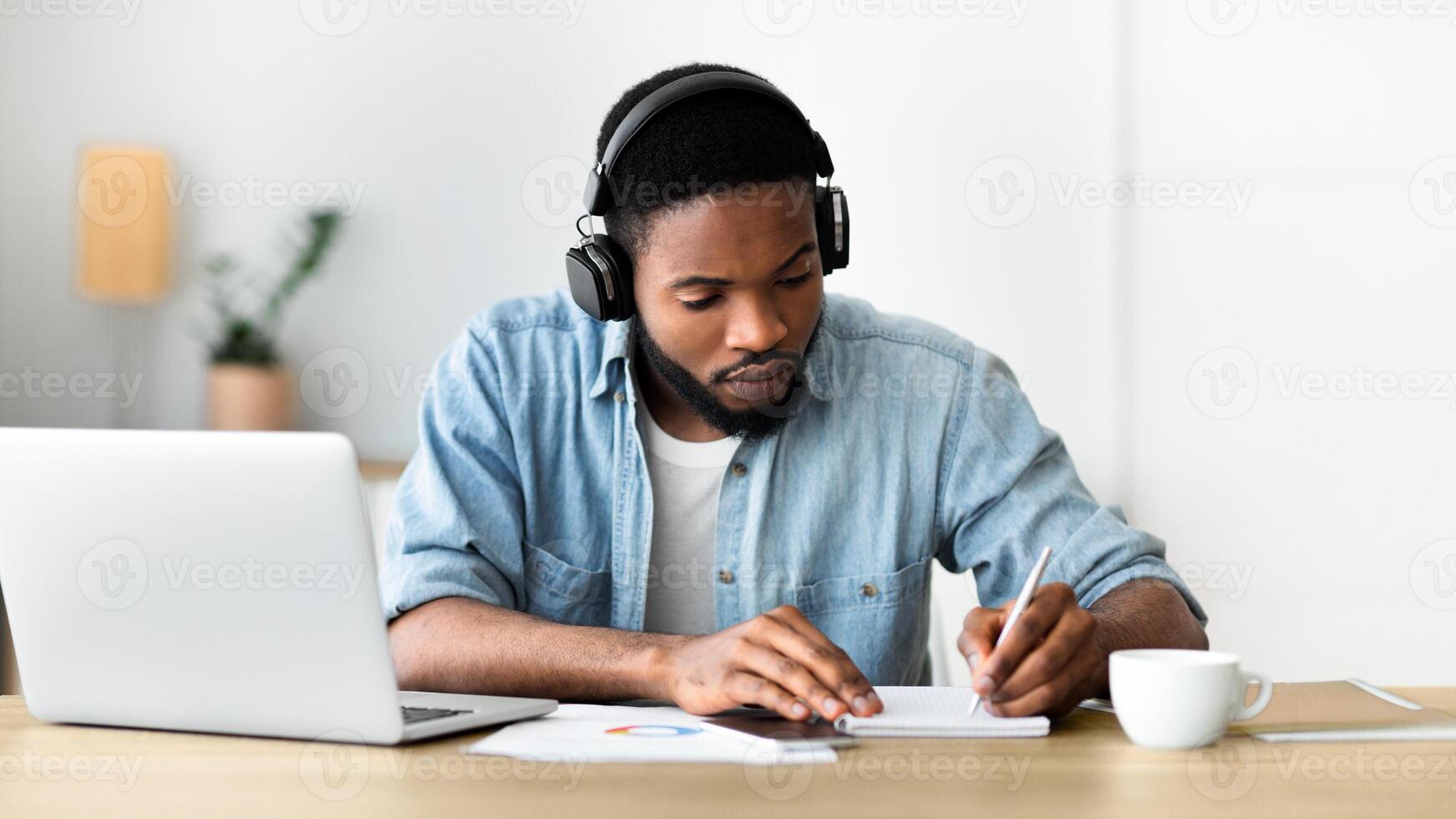 Concentrated african american guy in headphones studying foreign language online through conference and making notes in notebook, panorama with copy space photo
