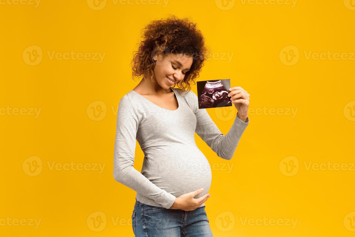 Happy afro pregnant woman with her baby sonography, enjoying first photo of her unborn child, pink background