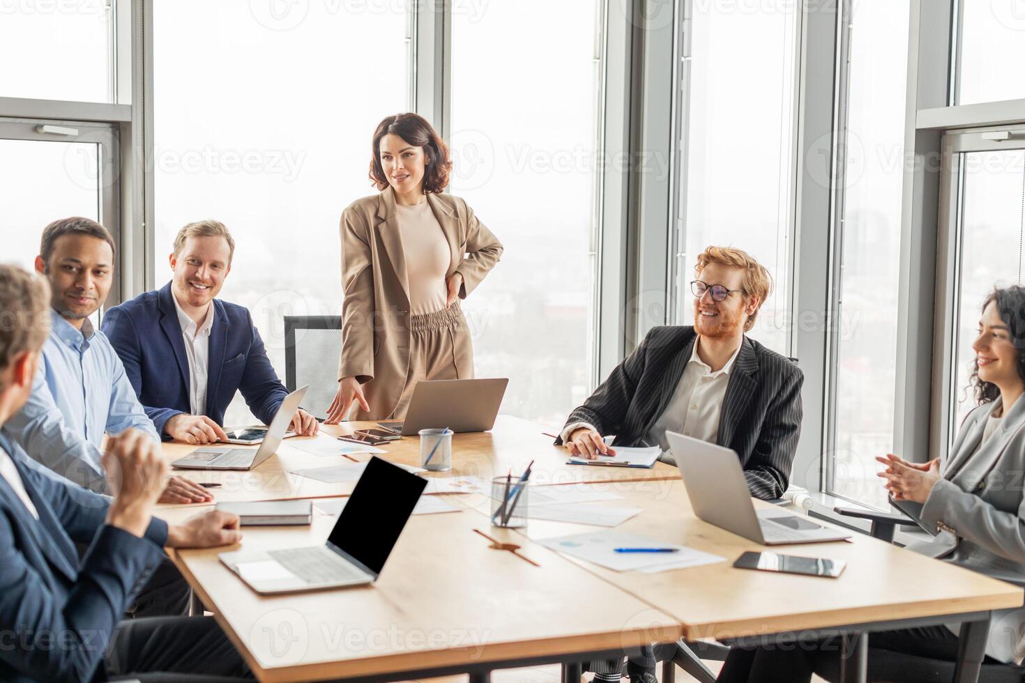 A diverse team of business professionals are gathered around a conference table in a modern office, engaging in a lively discussion. The woman standing at the head of the table, leading the meeting photo