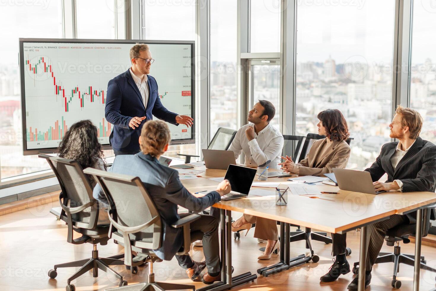 A businessman is presenting financial data to a team in a modern office with large windows overlooking the city. He is standing and pointing at a chart on a screen photo