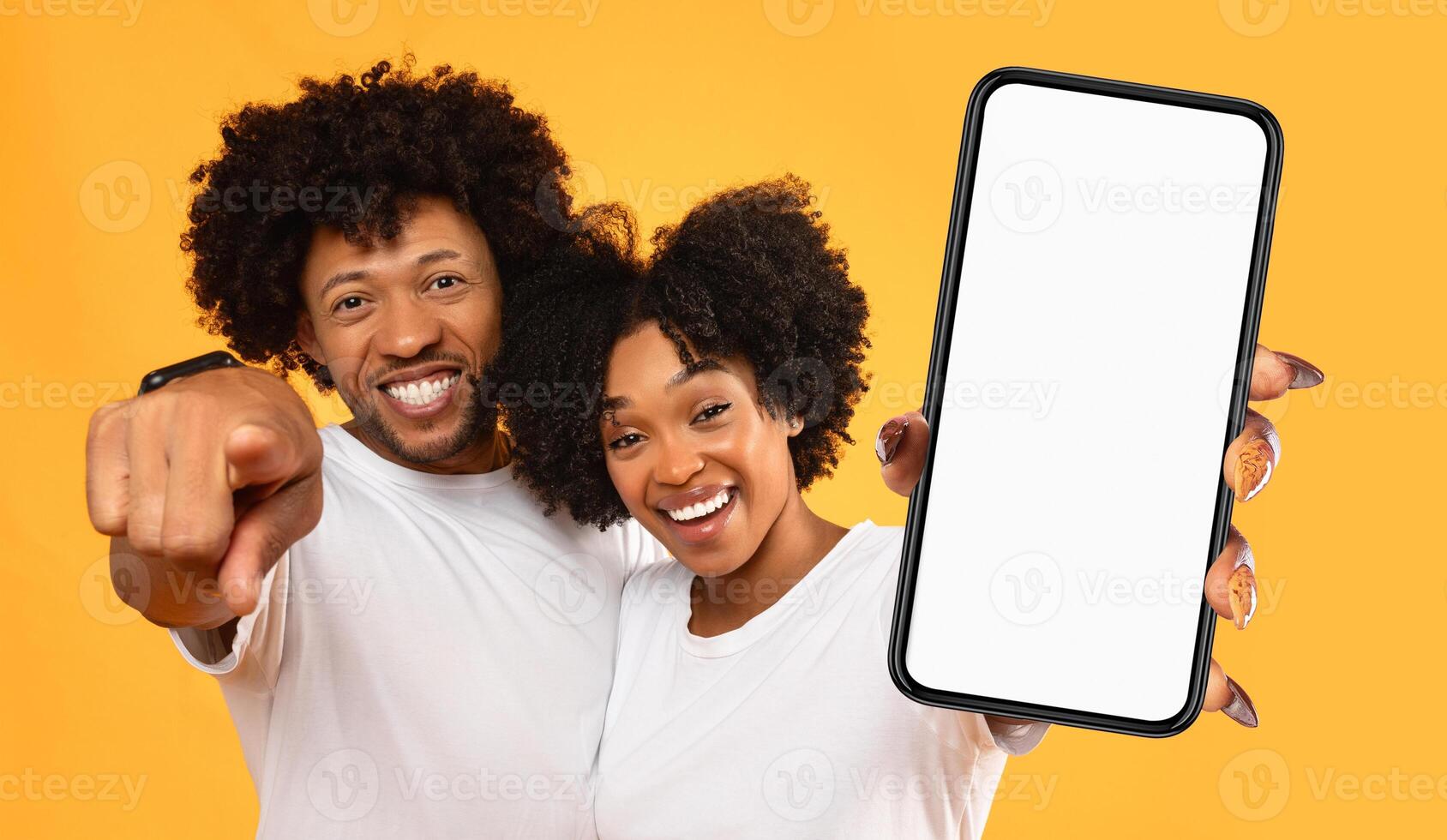 A young black couple with big smiles stand in front of a yellow background, holding a smartphone with a blank screen. The image is perfect for displaying a mobile app, website, or product. photo