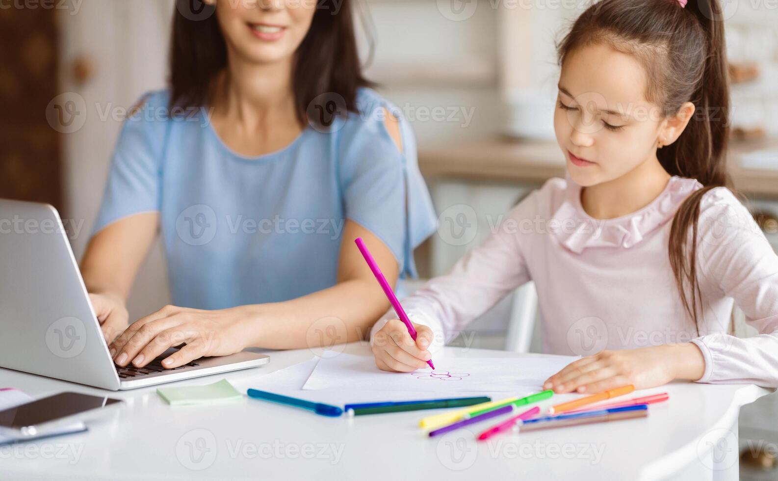 A woman sits at a table working on her laptop while her young daughter colors with markers. She is focused on her coloring and seems to be enjoying herself. photo