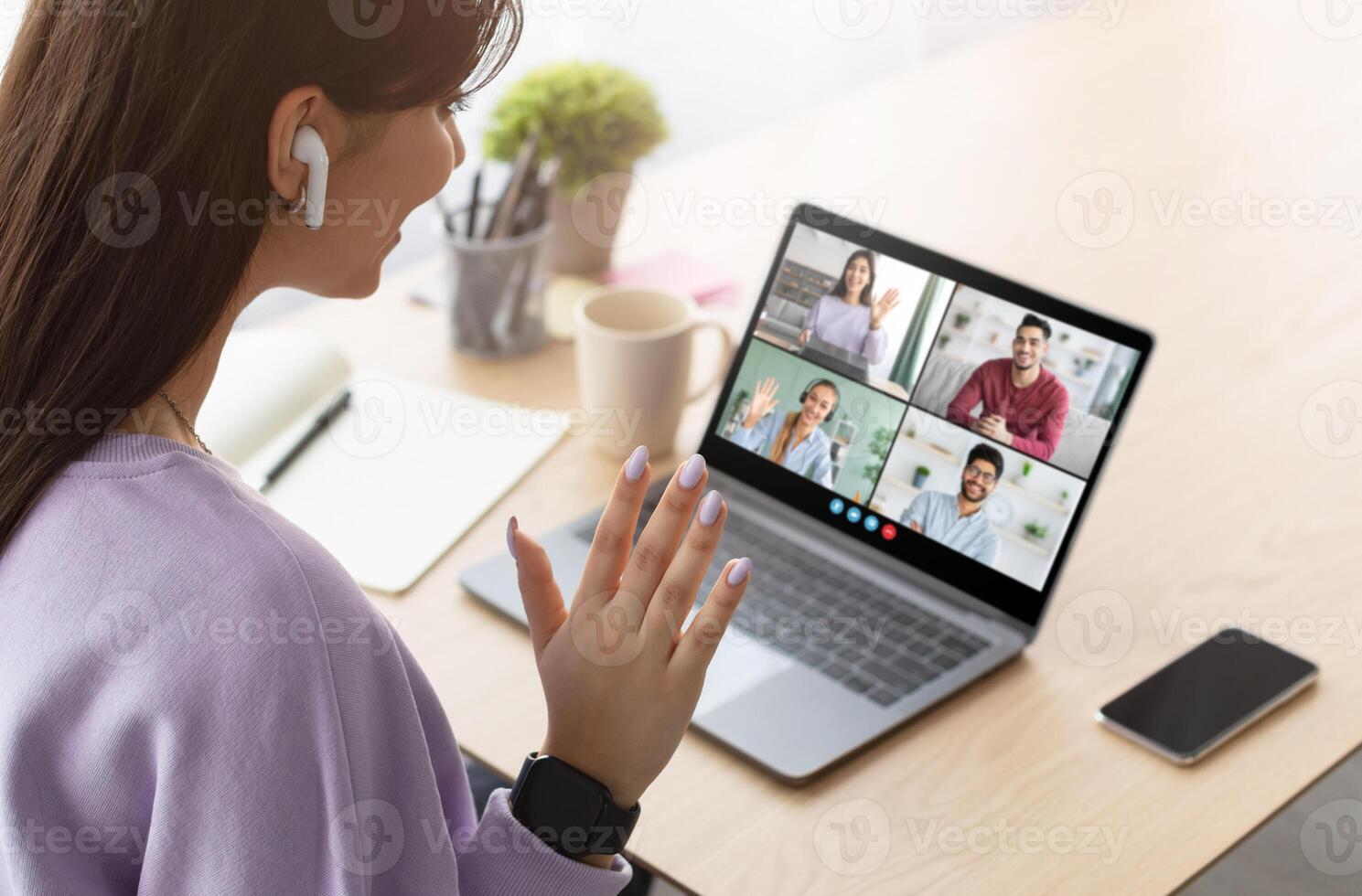 This image shows a young woman participating in a conference meeting at home. She is wearing a purple sweatshirt and has her hand raised to the camera, as if she is about to speak. photo