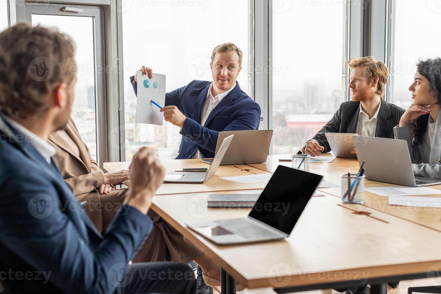 A group of business professionals are gathered around a table in an office. One man is presenting data from a document to the group while the other colleagues listen attentively, engage in discussion. photo