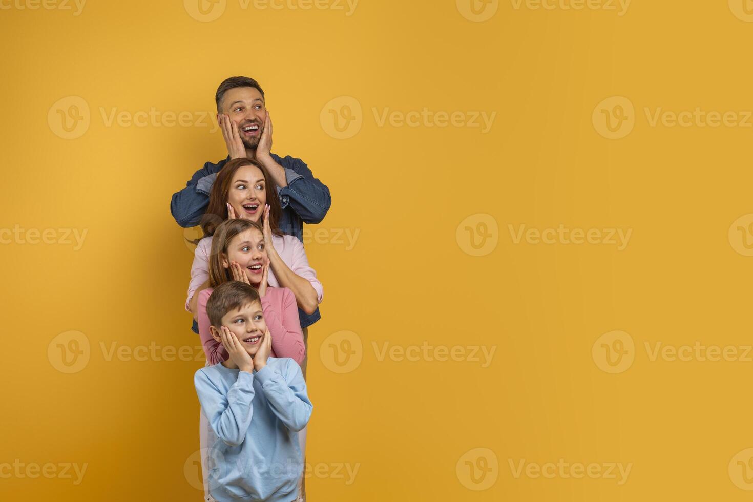 Surprised family of four, with two children and their parents, is lined up vertically, each placing their hands on their cheeks in a playful expression of surprise and happiness, copy space photo
