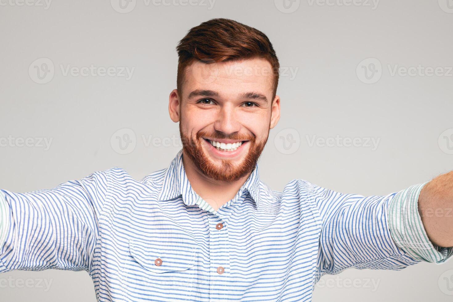 Cheerful handsome bearded redhaired man holding camera and making selfie on beige background, closeup photo