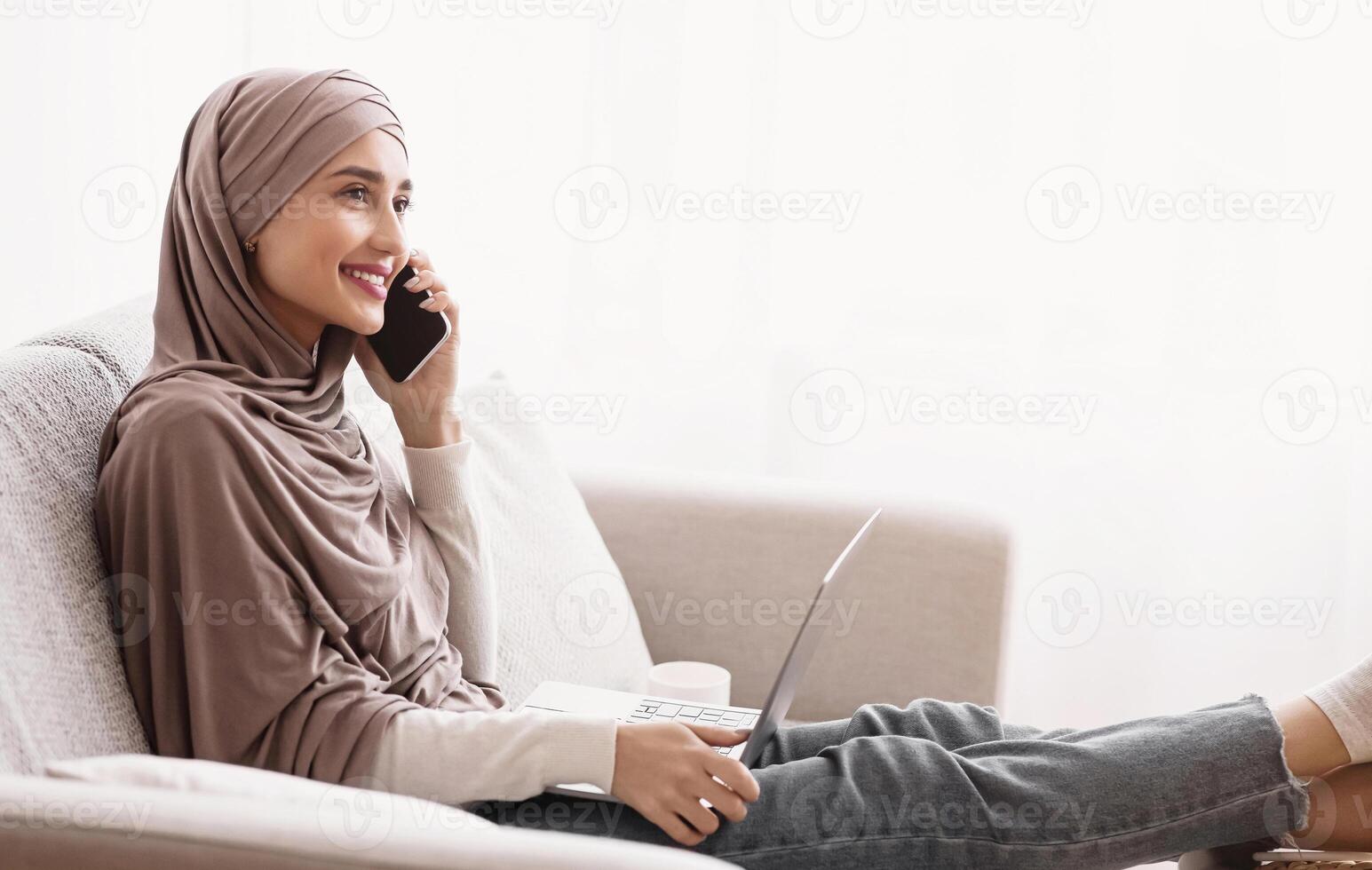Smiling Muslim Girl In Hijab Having Phone Conversation And Using Laptop While Sitting On Sofa Against Window At Home, Free Space photo