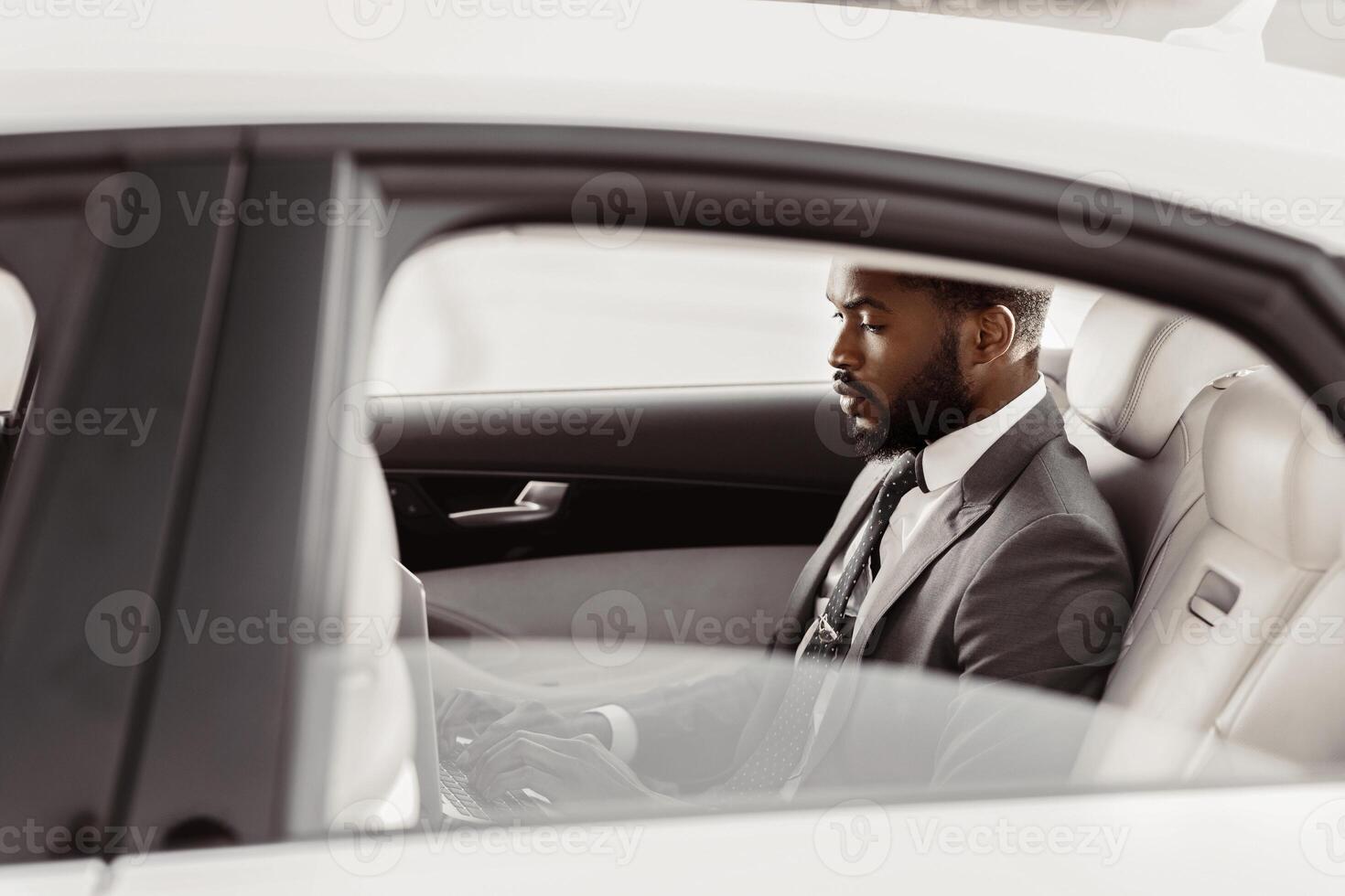 Black man in a suit is sitting in the back seat of a white car. He is looking down and appears to be thinking or working on something. The car is parked in a bright location, likely outdoors. photo