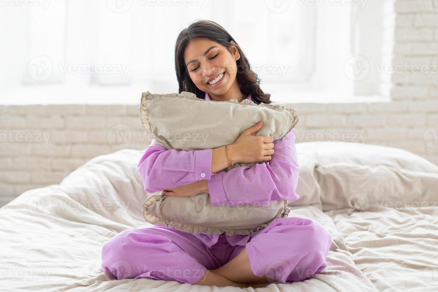 Happy middle eastern woman is seated on a bed, holding a pillow in her hands. She appears contemplative and relaxed in her bedroom setting. photo