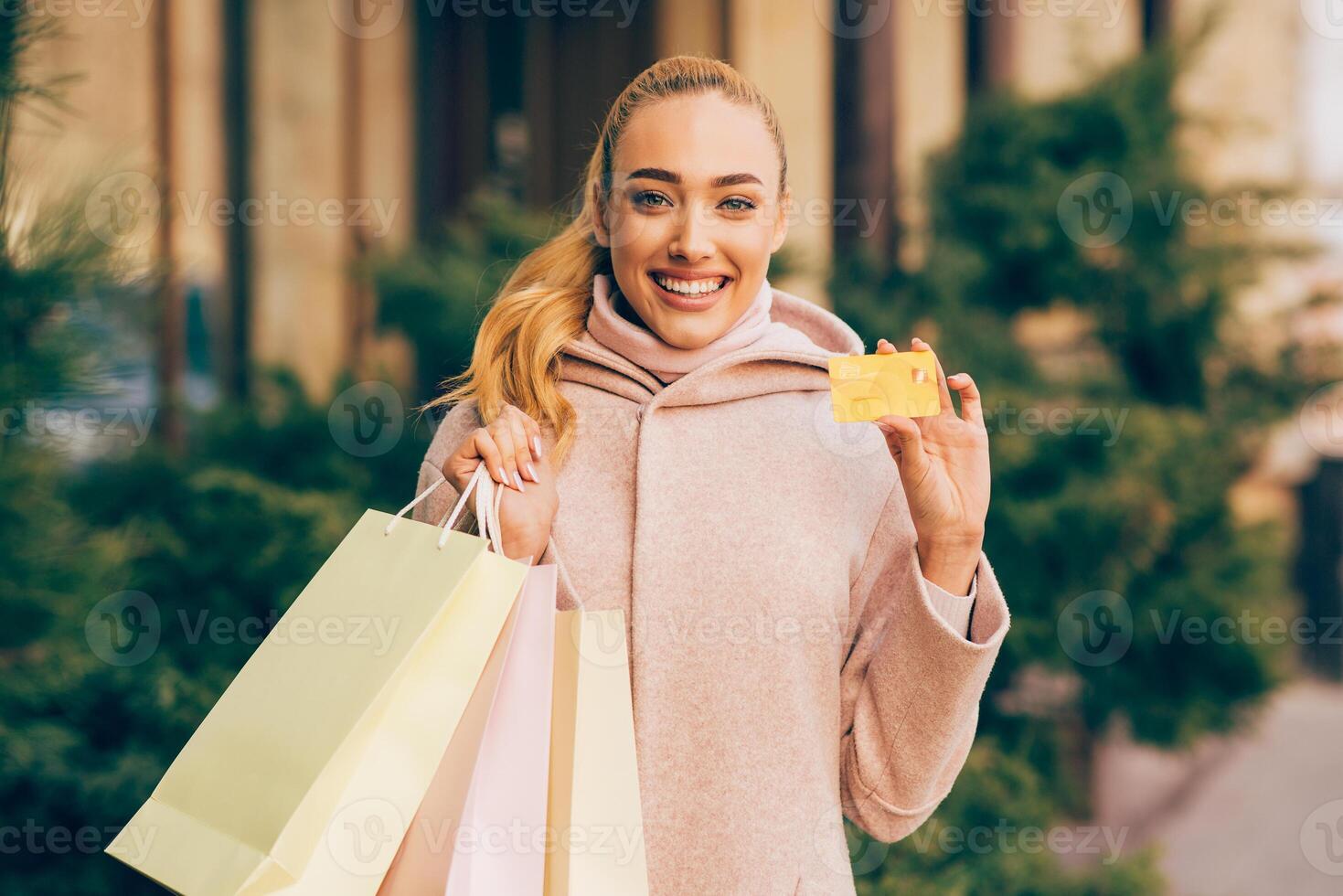 A young woman in a pink coat smiles and holds a credit card in her right hand, while holding several shopping bags in her left hand. She appears to be standing outside in a public area photo