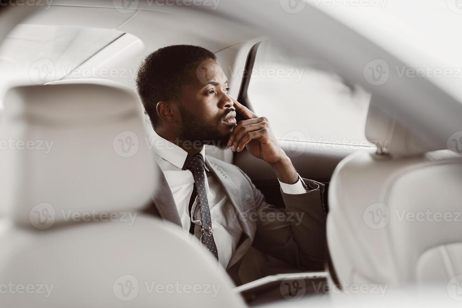An African American businessman sits in the back seat of a car, wearing a suit and tie. He is looking out the window, lost in thought. He has a hand on his chin photo