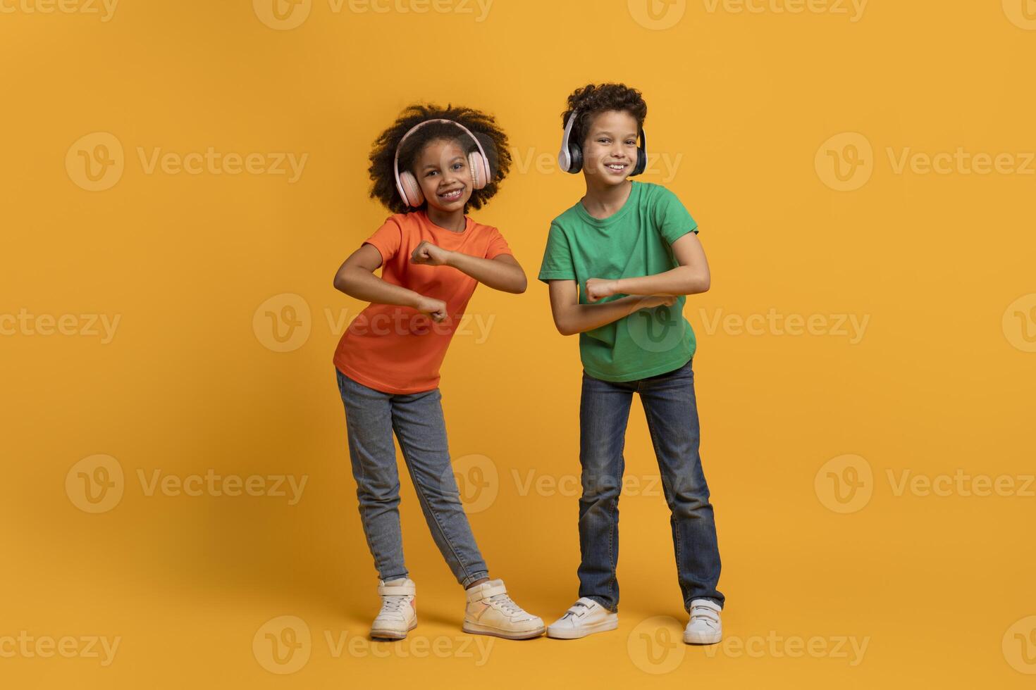 African American young boy and girl stand side by side, each striking a playful pose with their arms while dancing, using wireless headphones, listening to music photo