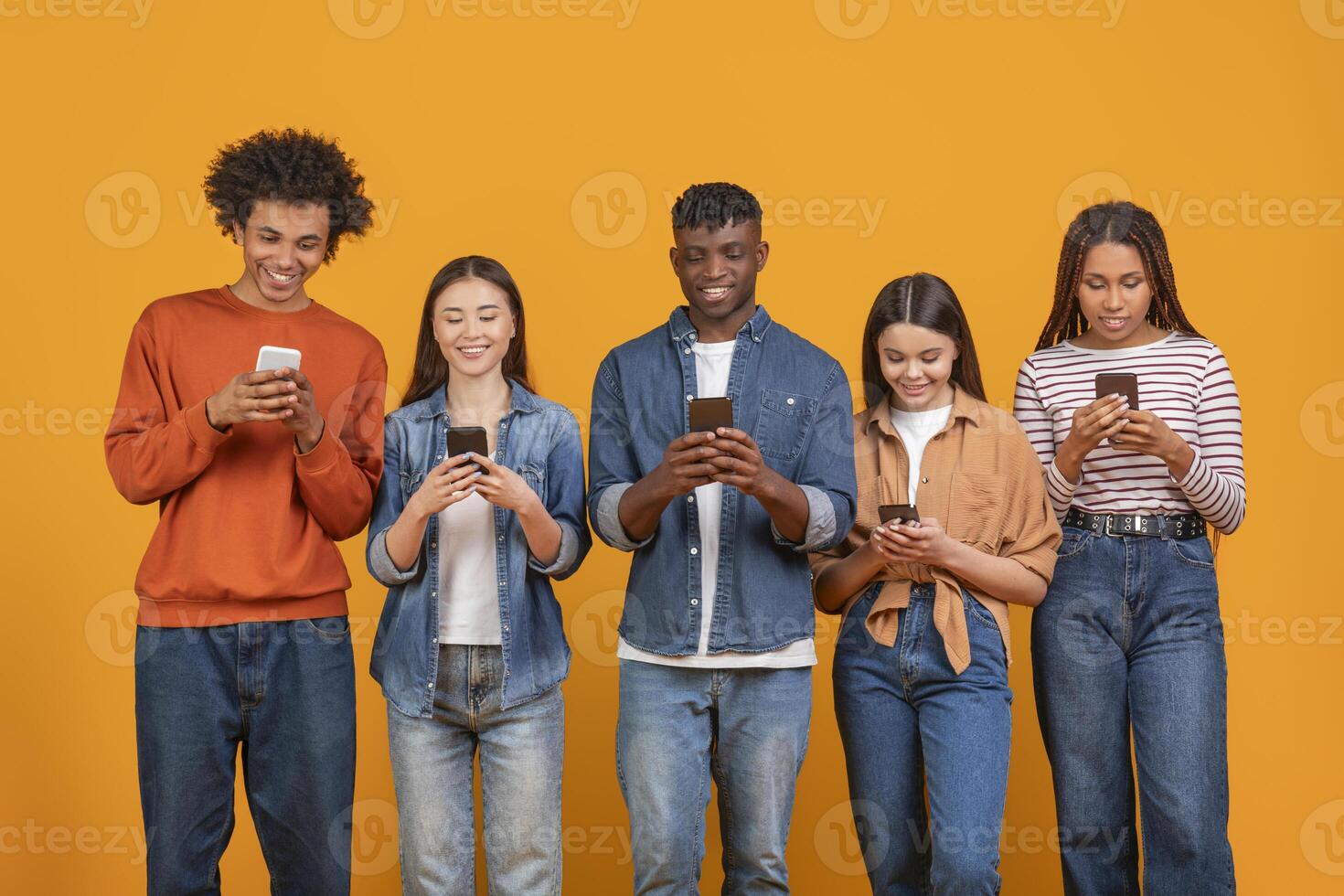 This image showcases a diverse group of five young adults, all using their smartphones while standing against a vibrant yellow background. They appear casual and relaxed photo