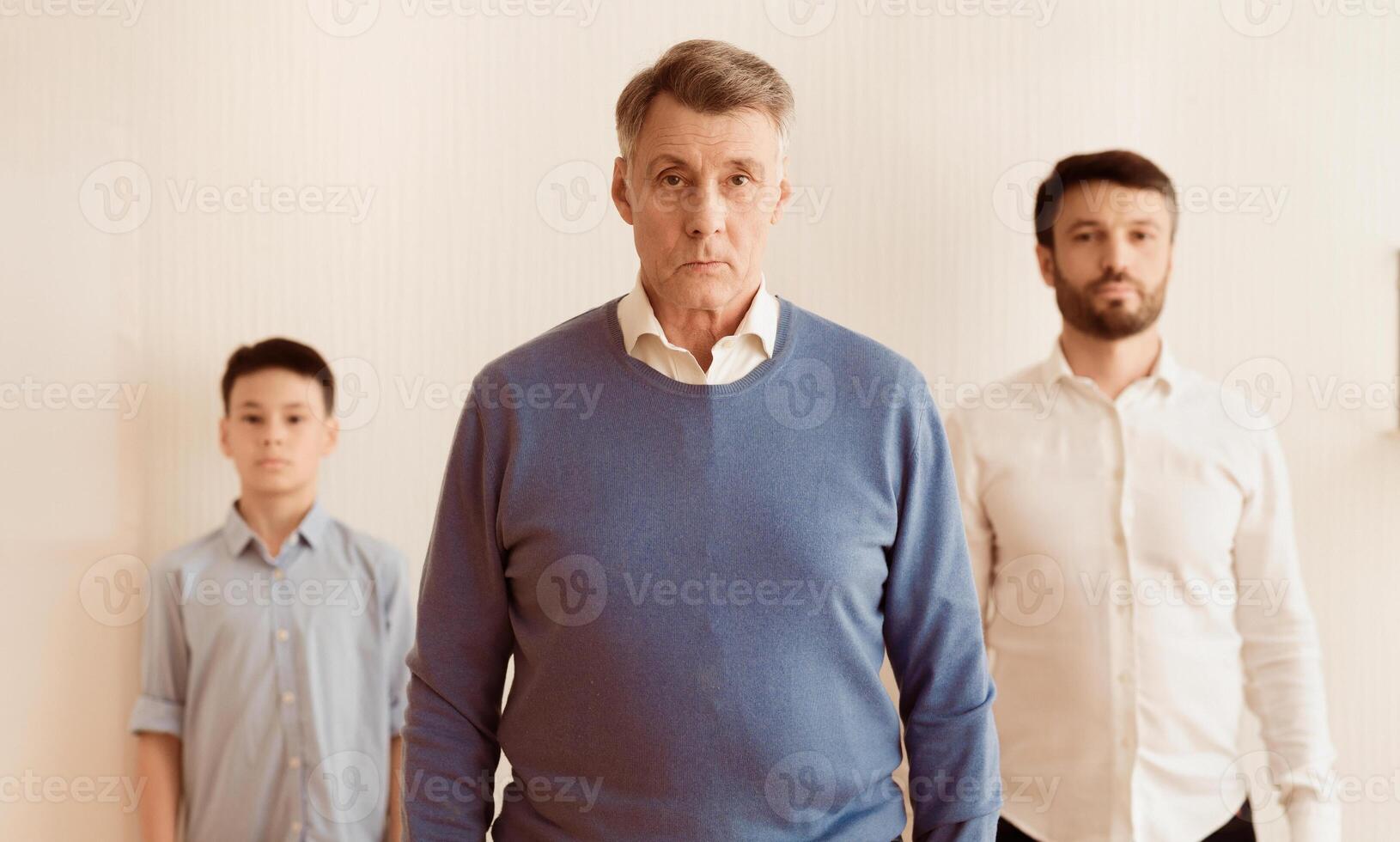 Serious Grandfather With Son And Grandson Looking At Camera Standing Against White Wall Indoor. Selective Focus photo