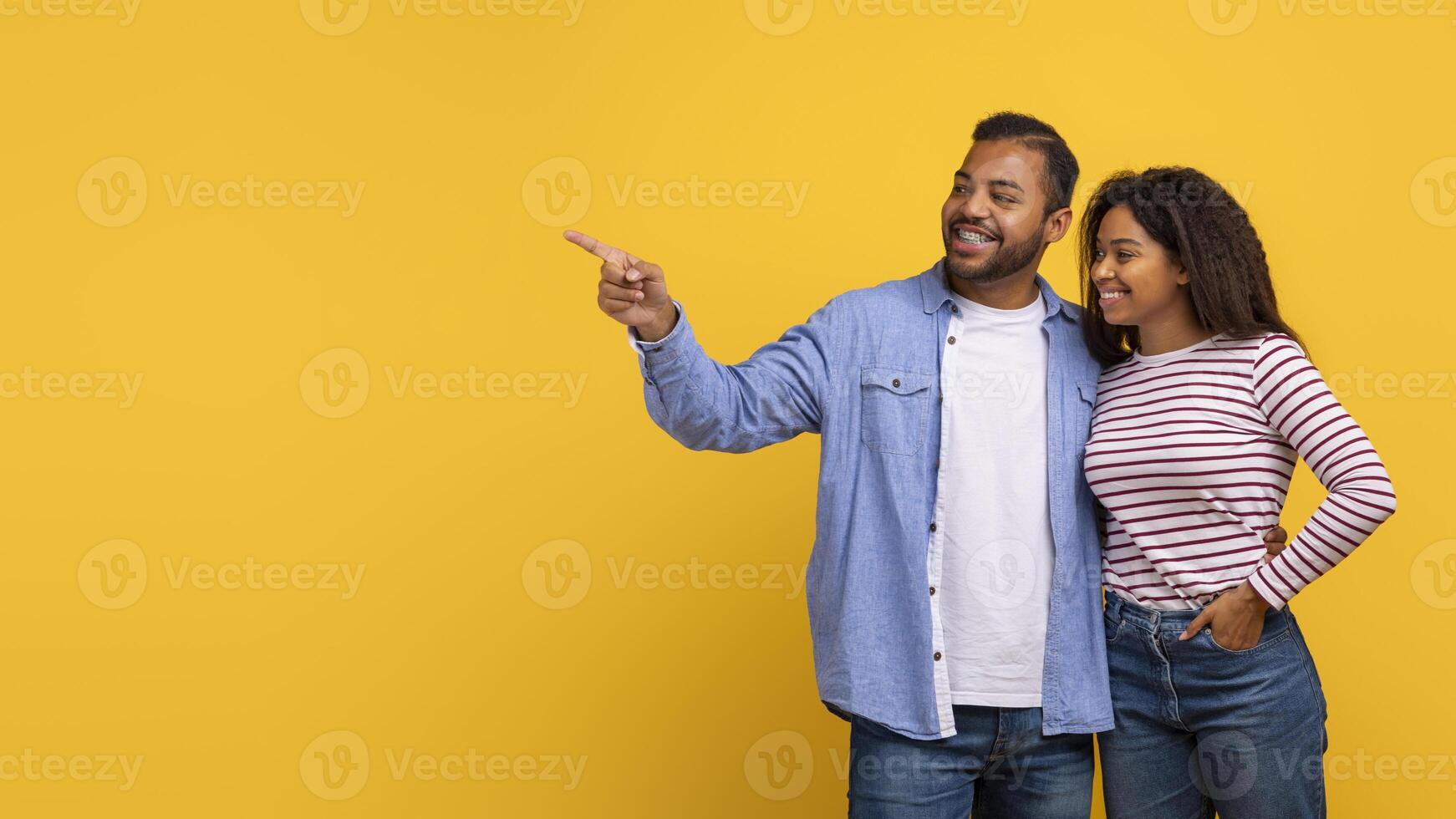 African American couple pointing at an unseen object while standing against a vibrant yellow background. They both appear engaged and focused on whatever they are indicating, copy space photo
