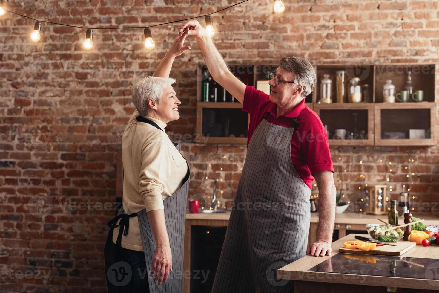 A senior couple, dressed in aprons, is laughing and dancing in their kitchen. There are string lights strung across the ceiling. The man is holding the woman hands as they sway photo
