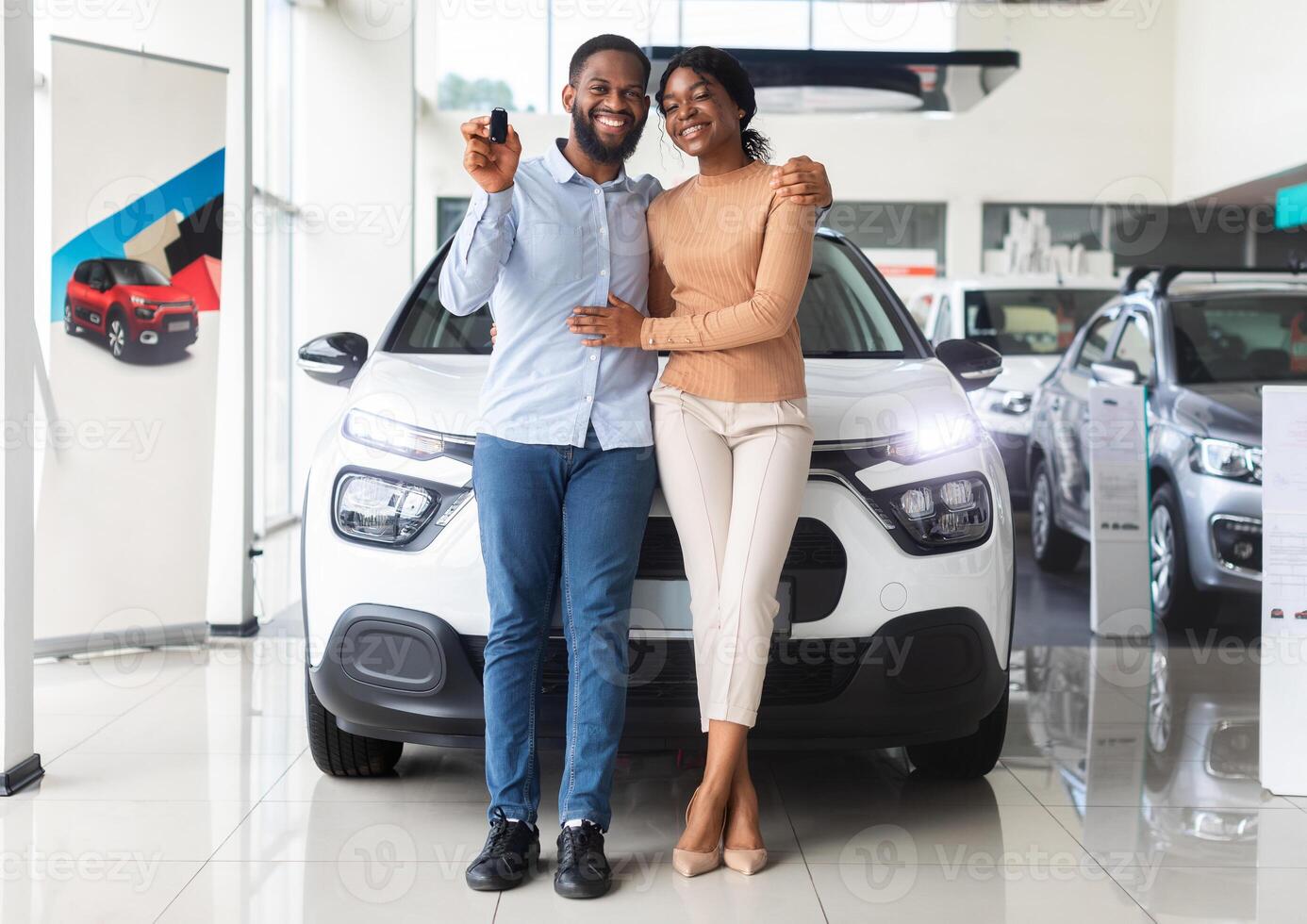 Beautiful african american couple bought car in dealership center, happy black spouses posing with keys and embracing, celebrating purchasing new family vehicle in automobile showroom, free space photo
