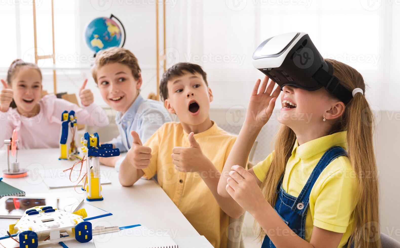 A group of elementary school students are engaged in a STEM class, with one student wearing a VR headset and experiencing a virtual world. The other students are looking on with excitement photo