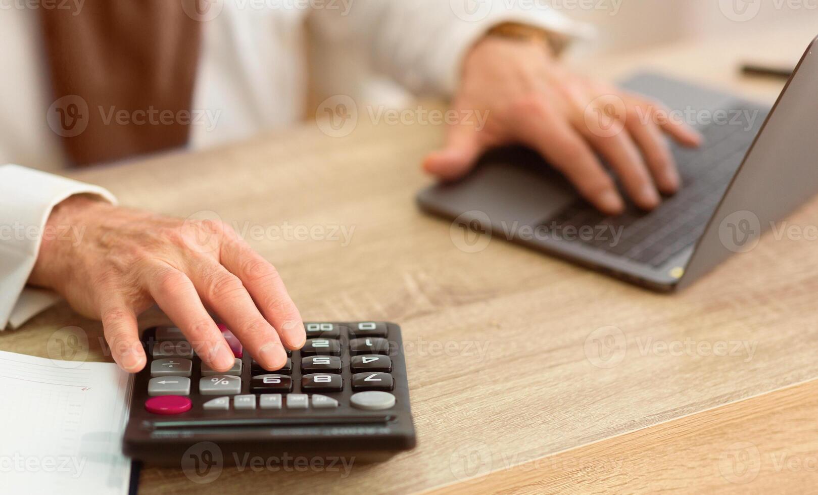 Closeup Of Elderly Man's Hands Calculating Finances Using Calculator And Laptop Sitting In Office. Cropped photo