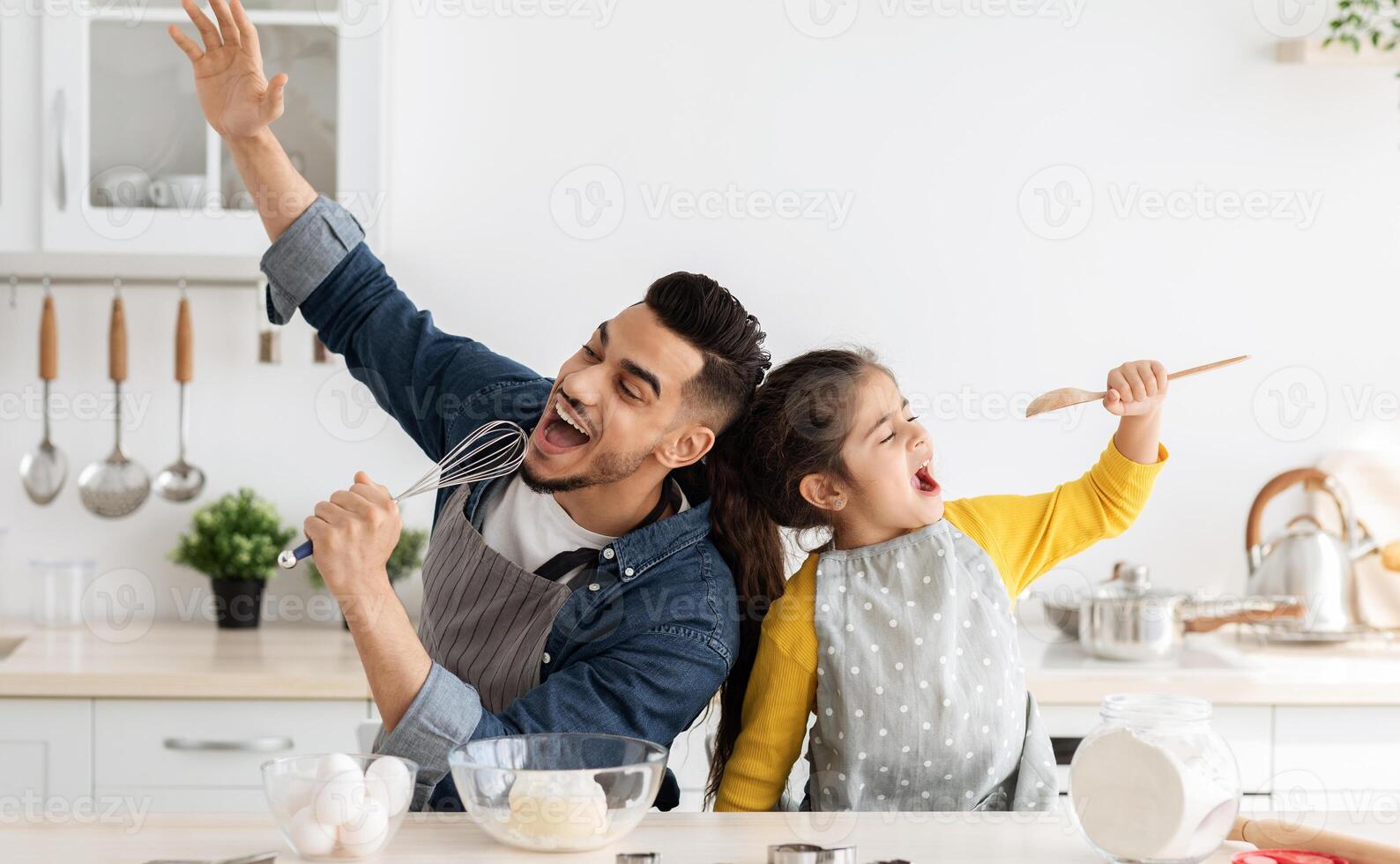 Cheerful arab father and little daughter having fun while baking together in kitchen, happy middle eastern dad and child singing and fooling, using spatula and whisk as microphones, copy space photo