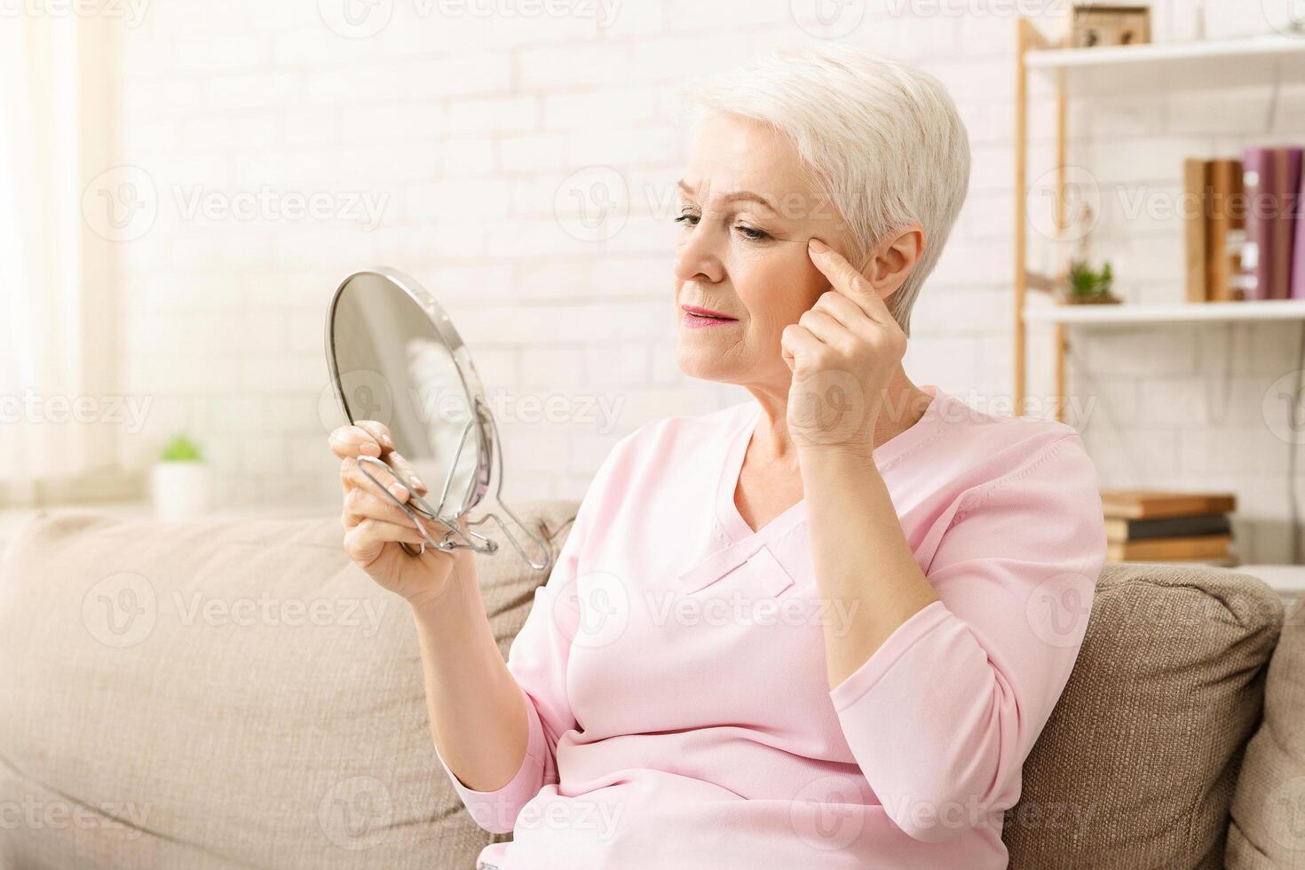 Elderly woman is seated on a couch, her gaze fixed on a mirror in front of her. She appears focused as she observes her reflection, possibly engaging in grooming or self-reflection. photo