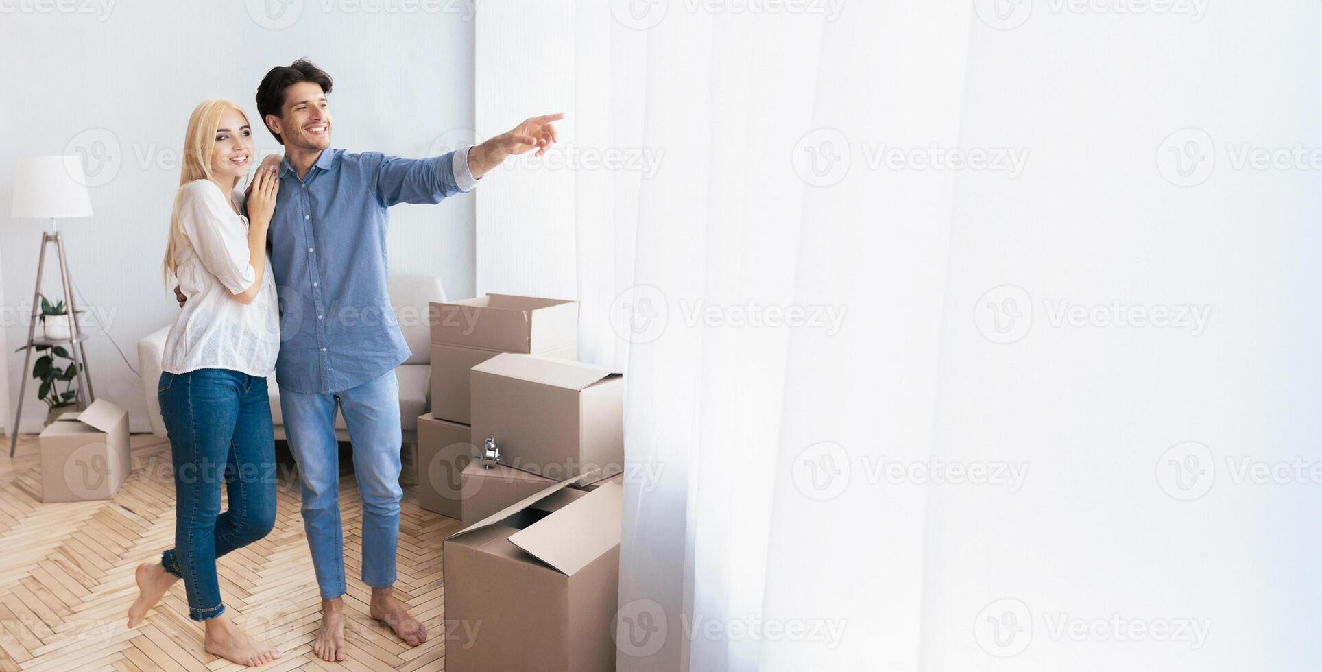 A young couple stands in their new home, looking out a window as they are surrounded by moving boxes. photo